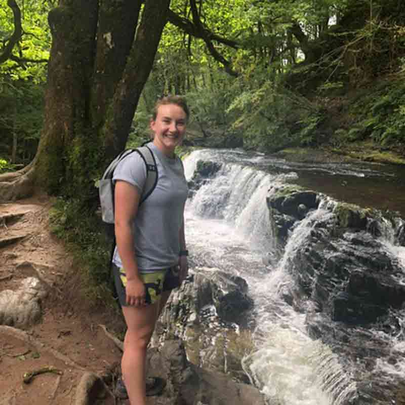 Portrait head shot photo of Maria Roynon wearing hiking attire in a forest area next to a water fall