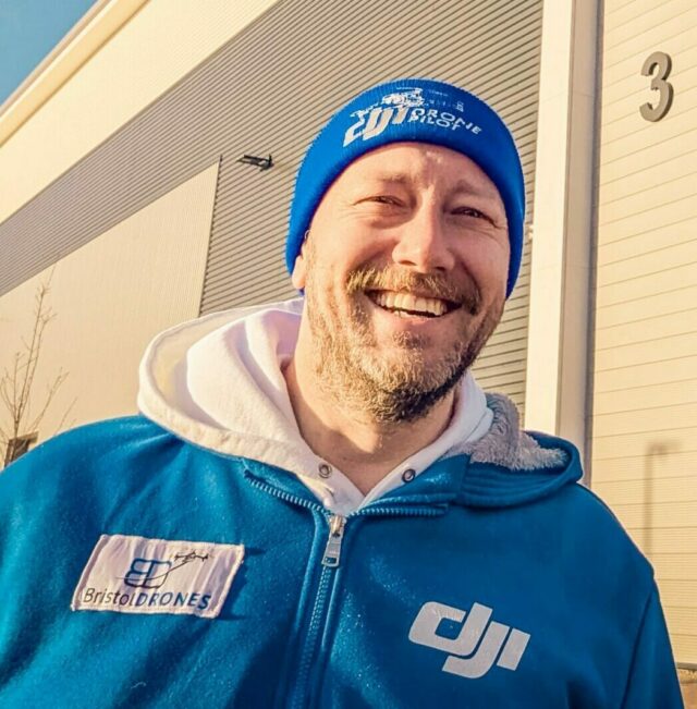 A smiling headshot of the found and drone operator, Philippe Francken, on a sunny winter day, wearing a blue fleece with the Bristol Drones logo stitched onto the left side and the DJI logo on the right, and a blue beanie hat saying Drone Pilot on it.